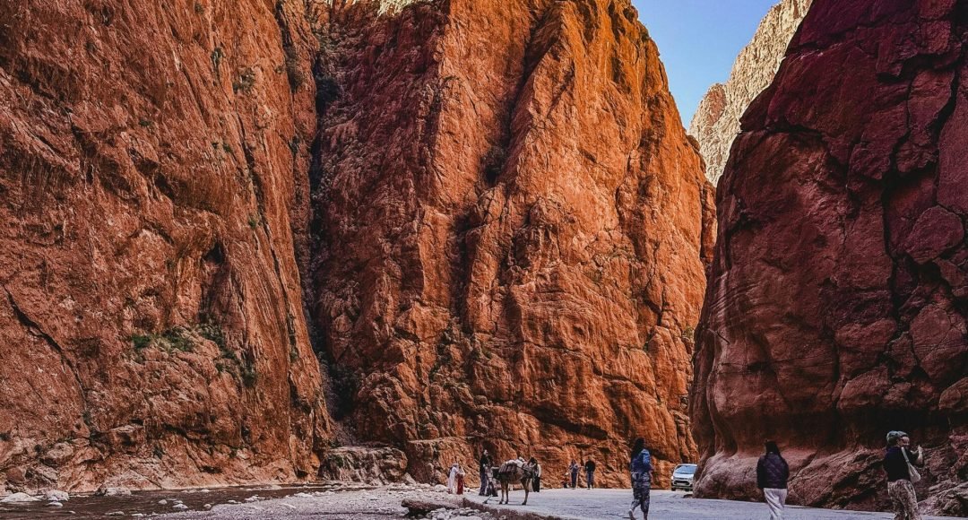 Sunlit red rock walls of Todra Gorge with people exploring the scenic path.